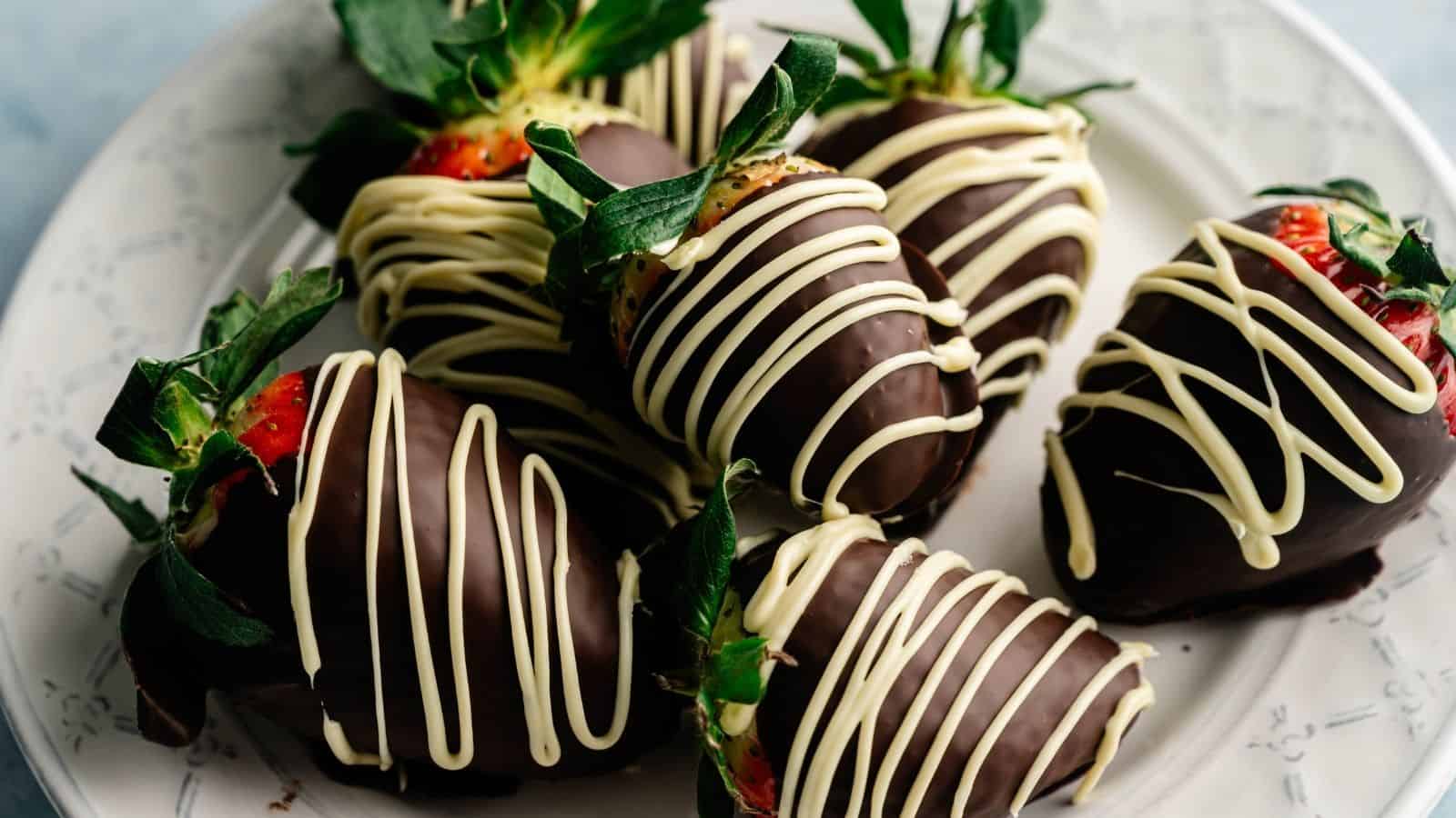 Six strawberries dipped in dark chocolate and drizzled with white chocolate are arranged on a white plate.