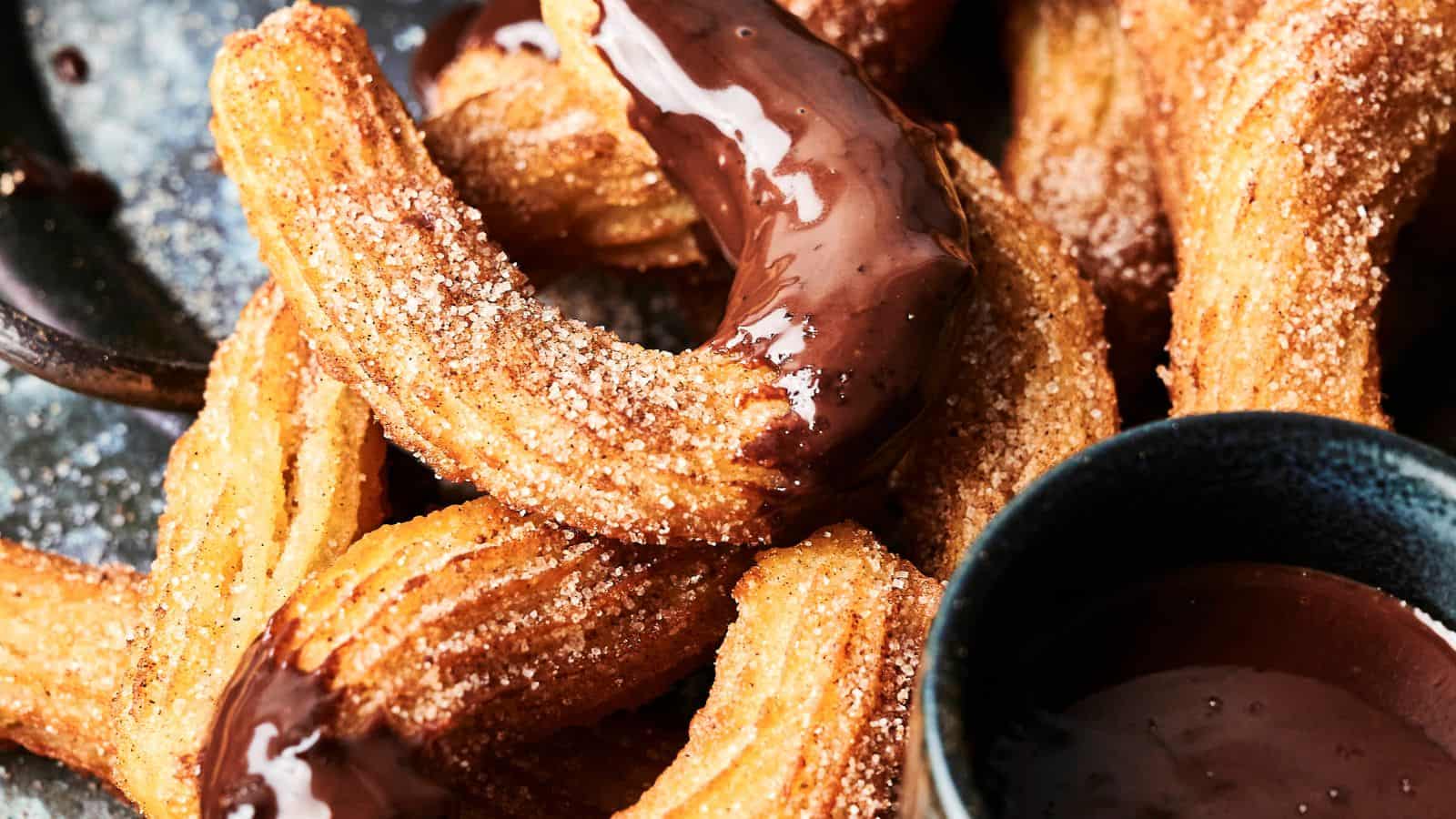 A close-up of churros coated in cinnamon sugar, some of which are dipped in chocolate sauce, with a bowl of chocolate sauce nearby.