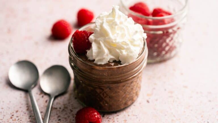 A small glass jar filled with chocolate mousse, topped with whipped cream and raspberries, sits next to two spoons and another jar of raspberries on a light surface.