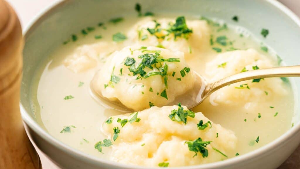A bowl of clear soup with several dumplings, garnished with chopped parsley, with a spoon lifting one dumpling.