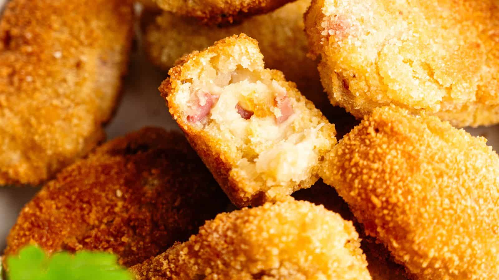Close-up of breaded and fried croquettes, with one cut open to reveal a creamy filling mixed with diced ham.