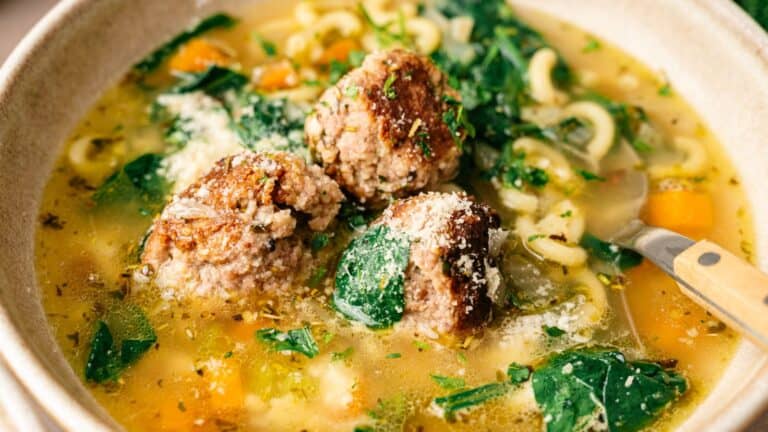 A bowl of soup with meatballs, pasta, spinach, carrots, and broth, garnished with grated cheese and herbs, with a spoon in the bowl.