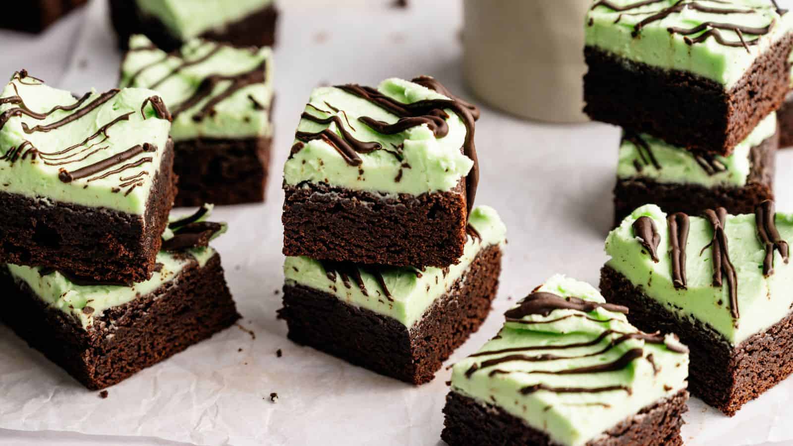 Brownie squares topped with a layer of green mint frosting and drizzled with chocolate, arranged on parchment paper.