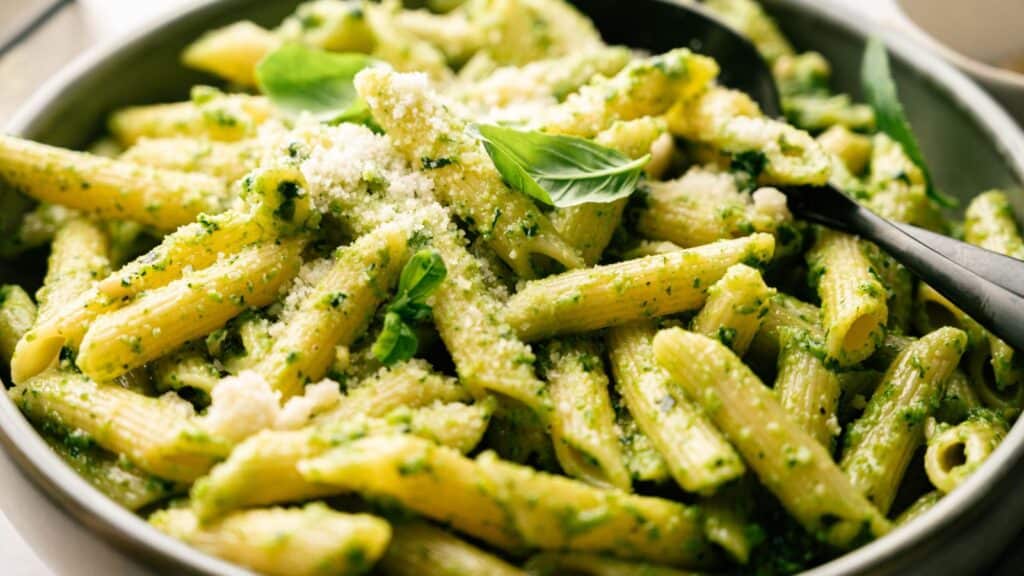 A bowl of penne pasta coated in green pesto sauce, garnished with grated parmesan cheese and fresh basil leaves, with a black serving spoon.
