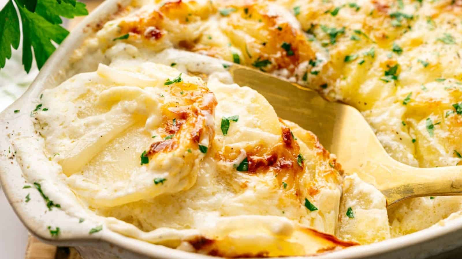 A close-up of creamy, baked scalloped potatoes with melted cheese, garnished with chopped parsley, in a white baking dish with a serving spoon.