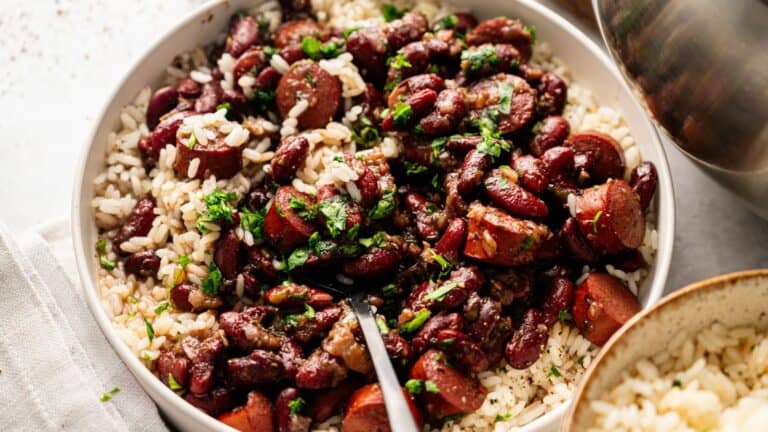 A bowl of white rice topped with red beans, sliced sausage, and chopped herbs, with a spoon resting inside.