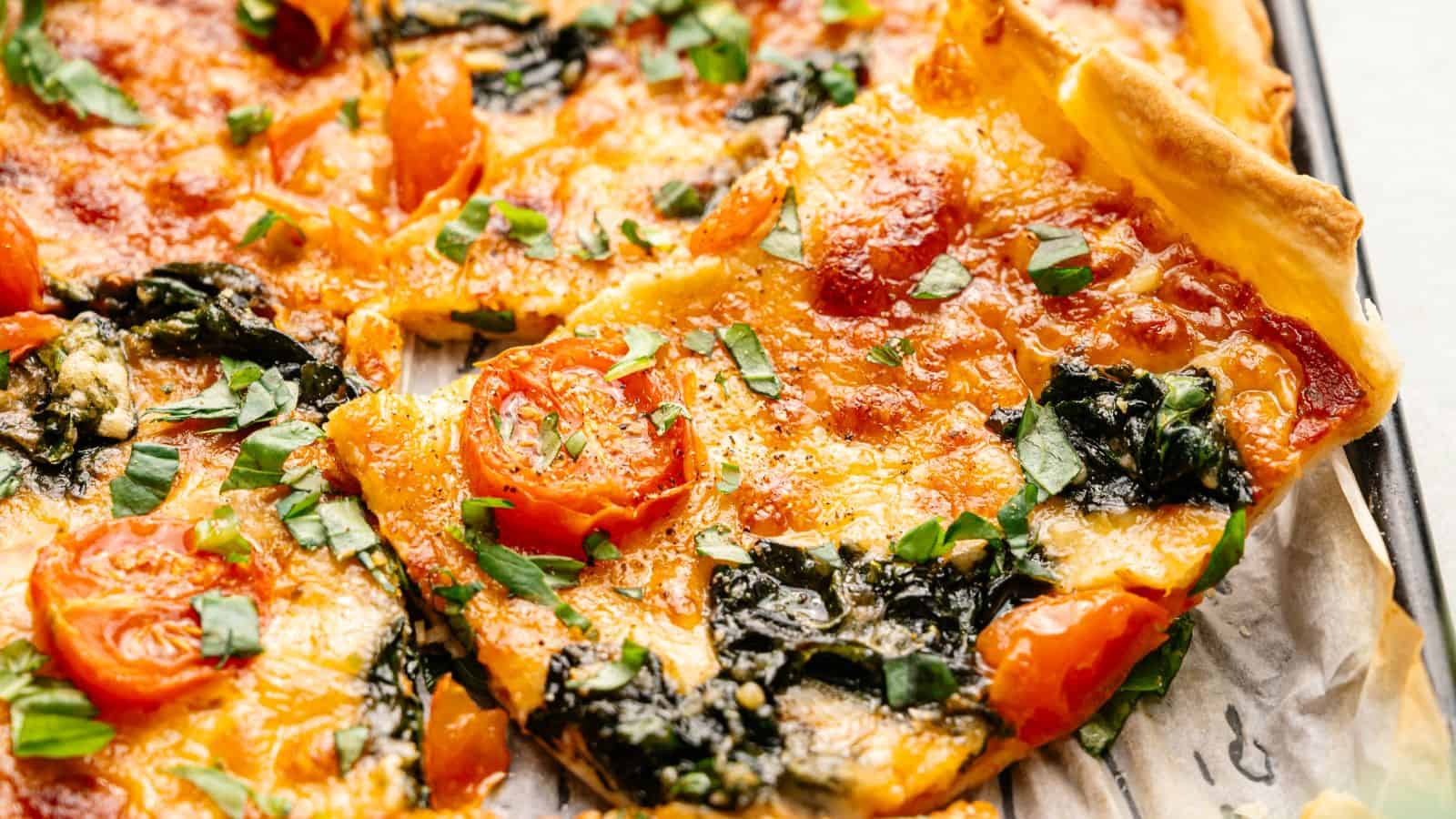A close-up of a rectangular pizza topped with melted cheese, cherry tomatoes, spinach, and fresh herbs, on a baking tray lined with parchment paper.