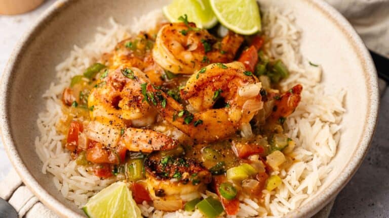 Bowl of rice topped with shrimp in a seasoned tomato and vegetable sauce, garnished with chopped herbs and lime wedges.
