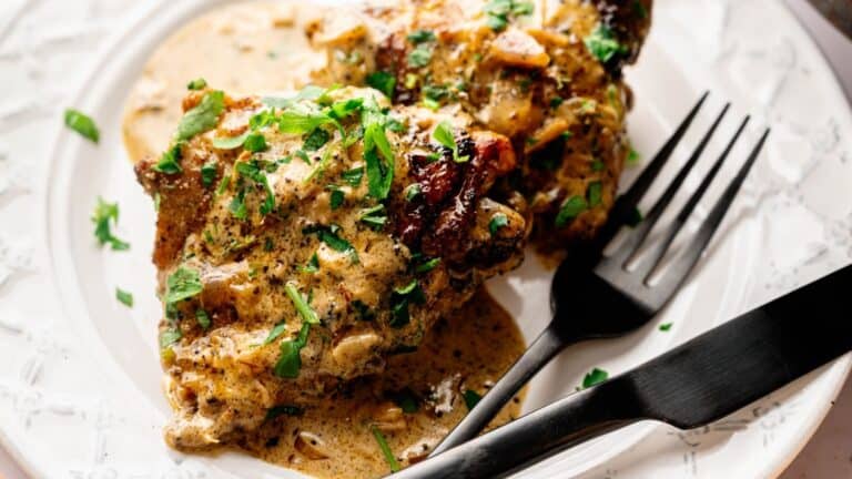 Cooked chicken pieces with creamy sauce and chopped herbs served on a white plate, accompanied by a black fork and knife.