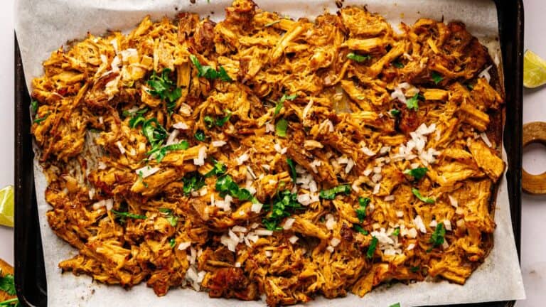 Shredded seasoned meat spread on a parchment-lined baking sheet, topped with chopped onions and fresh herbs. Lime wedges are visible at the corners.