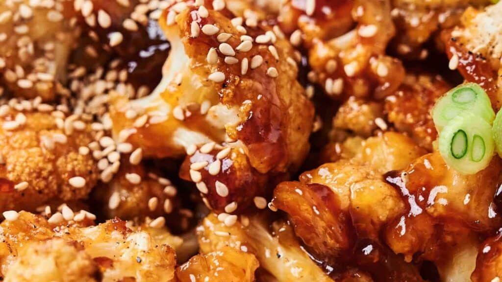 Close-up of roasted cauliflower florets coated in a dark sauce, topped with sesame seeds and garnished with sliced green onions.