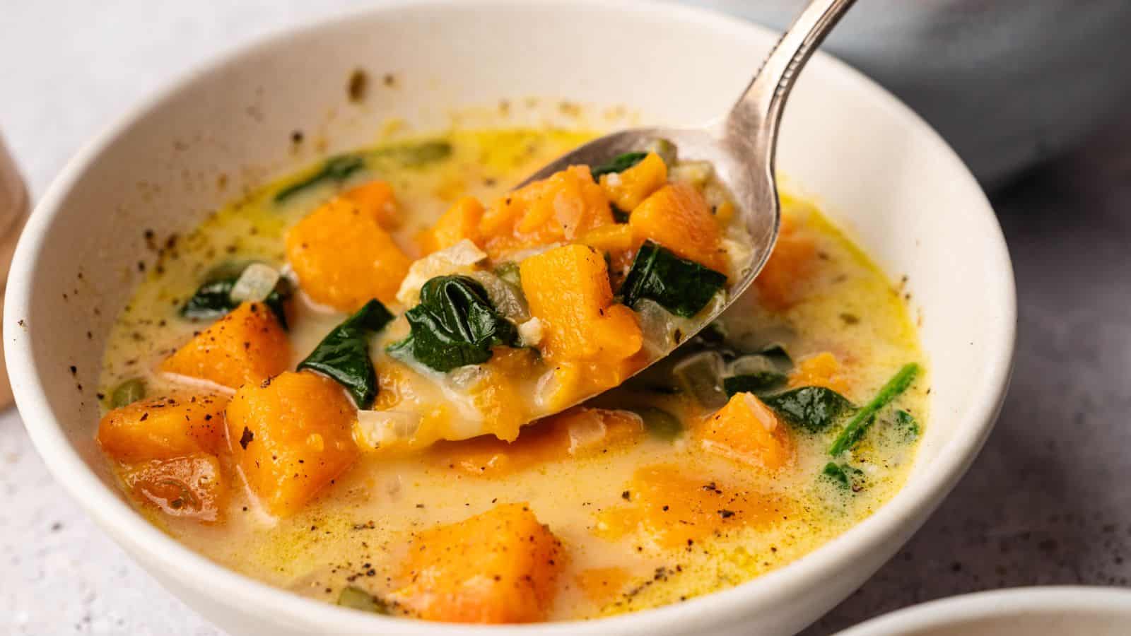 A bowl of creamy soup containing chunks of orange squash, leafy spinach, and visible seasoning, with a spoon lifting a portion.