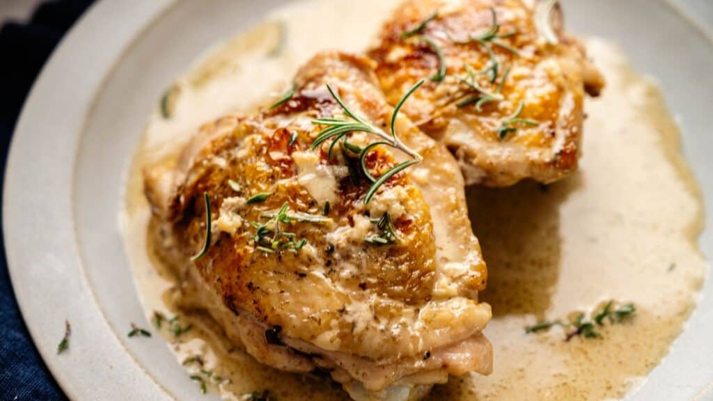 Two pieces of cooked chicken in a creamy sauce, garnished with fresh rosemary and thyme, served on a light-colored plate.