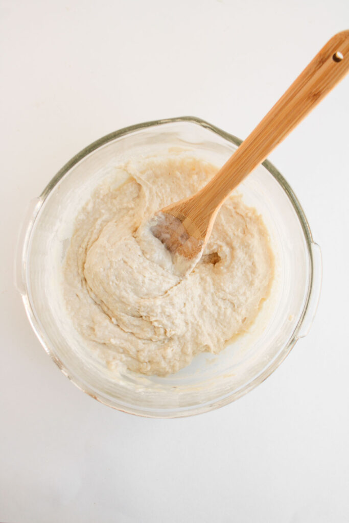 A glass bowl containing thick, pale batter for Muffin Pan Strawberry Shortcakes being mixed with a wooden spoon on a white surface.