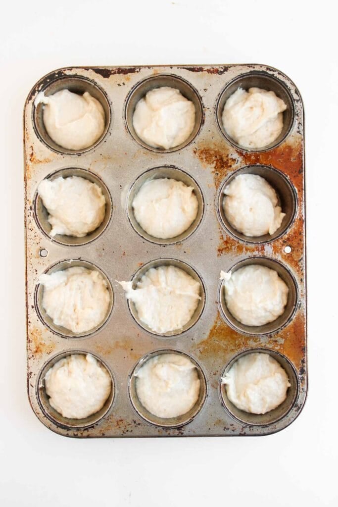 A 12-cup muffin pan filled with raw dough for Strawberry Shortcakes sits ready to be baked on a white background.