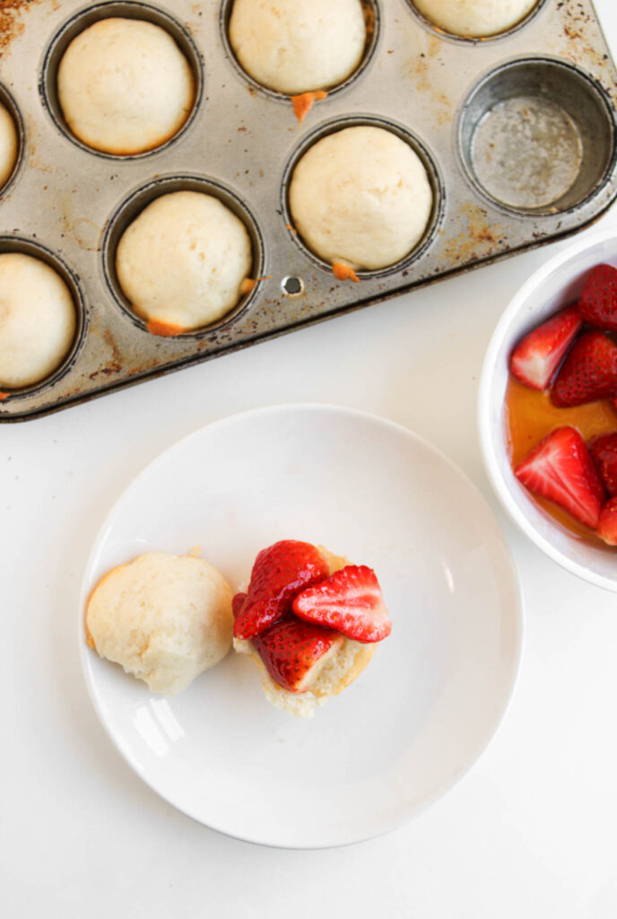 A muffin tin with baked Muffin Pan Strawberry Shortcakes, a white plate featuring a split shortcake generously topped with fresh strawberries, and a bowl filled with sliced strawberries.