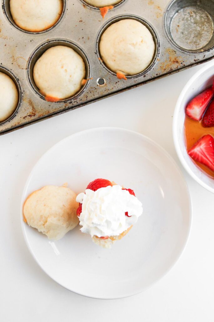 A muffin pan filled with perfectly baked Muffin Pan Strawberry Shortcakes, a plate featuring one split shortcake crowned with juicy strawberries and fluffy whipped cream, and a bowl of freshly sliced strawberries.