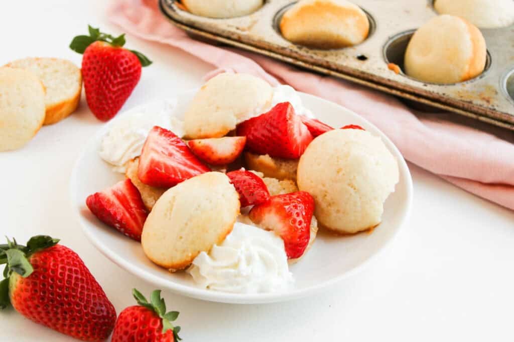 A plate with Muffin Pan Strawberry Shortcakes&mdash;shortcake biscuits, sliced strawberries, and whipped cream&mdash;sits beside whole strawberries and a muffin tin filled with extra biscuits.
