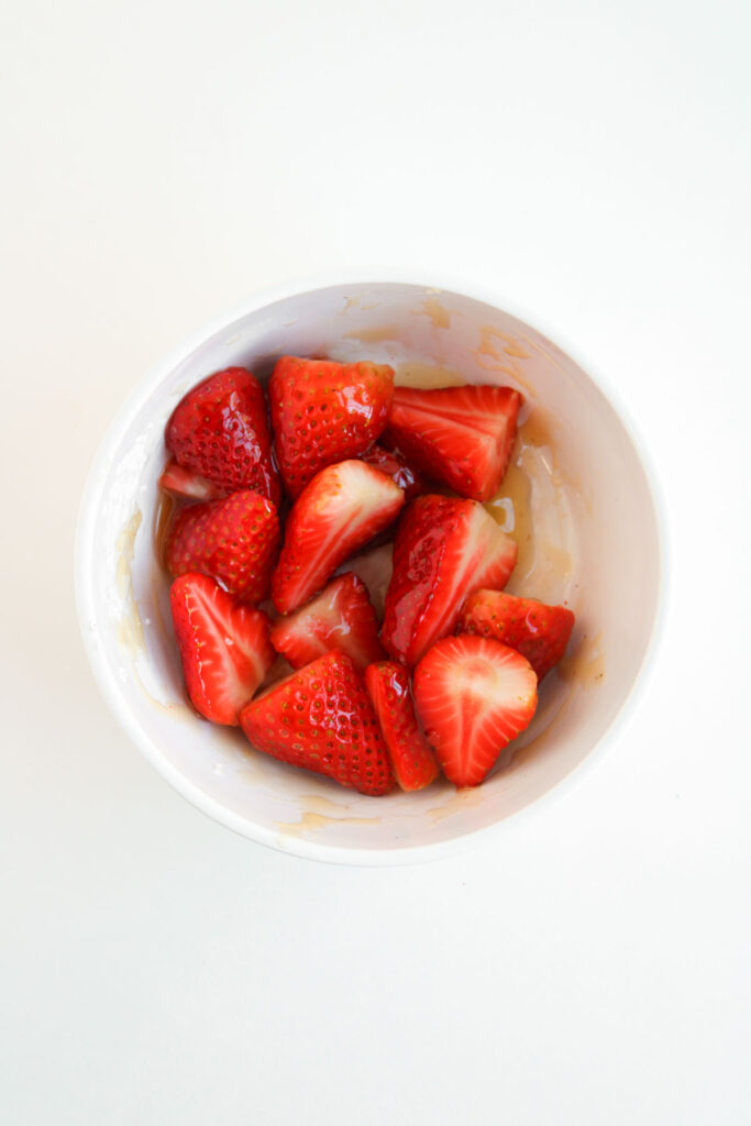 A white bowl filled with sliced strawberries coated in a light syrup, reminiscent of the sweet topping used for Muffin Pan Strawberry Shortcakes, viewed from above on a plain white background.