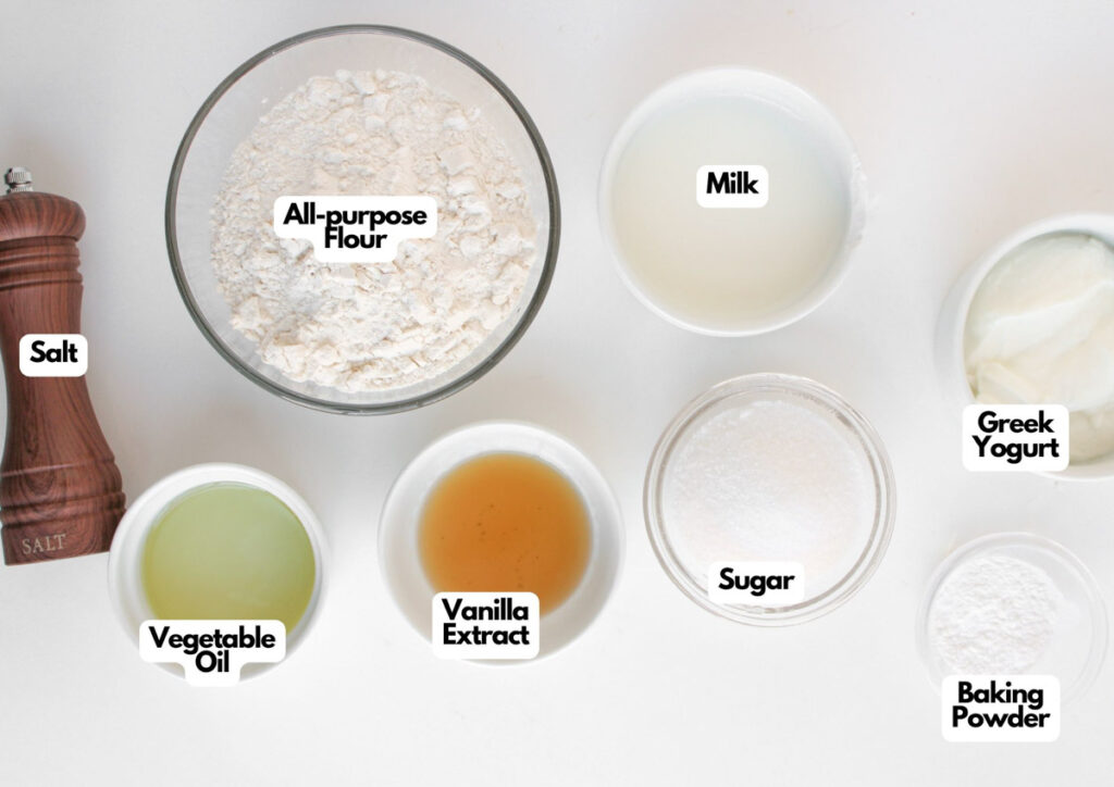 Seven labeled baking ingredients in small bowls—perfect for making Muffin Pan Strawberry Shortcakes: all-purpose flour, milk, Greek yogurt, baking powder, sugar, vanilla extract, vegetable oil, and a salt shaker.