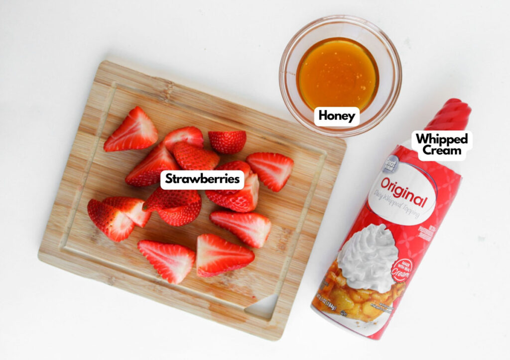 A small cutting board with sliced strawberries, a bowl of honey, and a can of whipped cream on a white surface—perfect ingredients for assembling delightful Muffin Pan Strawberry Shortcakes.