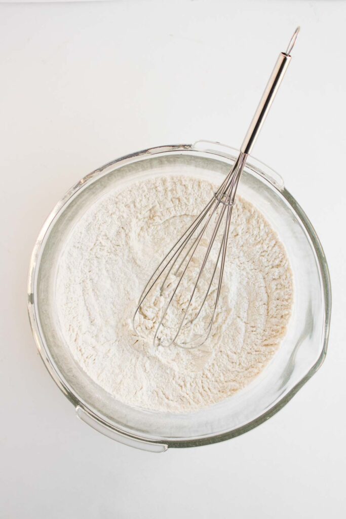 A metal whisk rests in a glass bowl filled with flour mixture on a white surface, ready to prep the batter for Muffin Pan Strawberry Shortcakes.