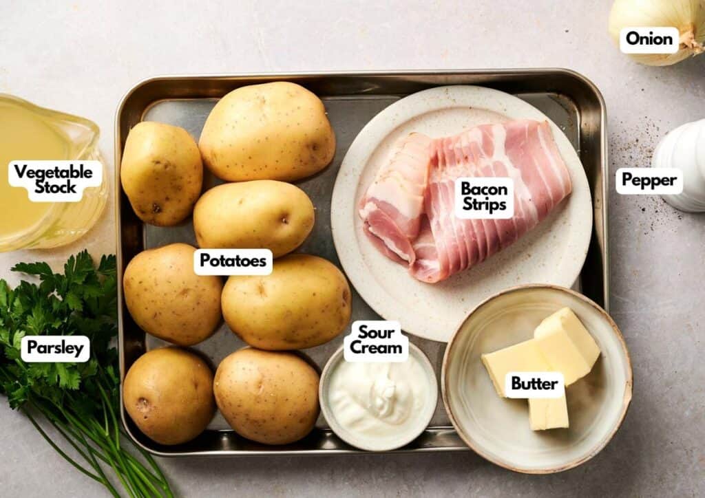 A tray with ingredients for Air Fryer Potato Soup: potatoes, bacon strips, sour cream, and butter, plus bowls of vegetable stock, parsley, pepper, and an onion. Each ingredient is clearly labeled.