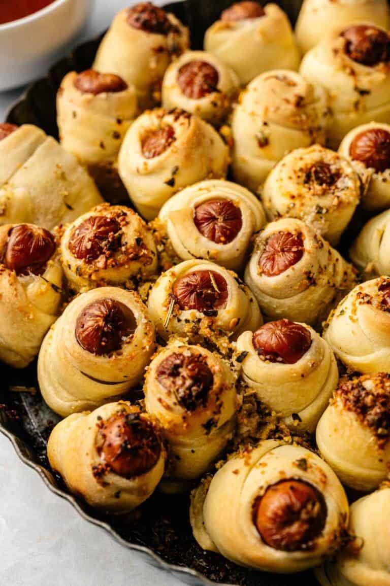 A tray of Pull-Apart Pigs in a Blanket, featuring mini sausages wrapped in golden-brown dough, sprinkled with herbs and grated cheese for a deliciously shareable treat.