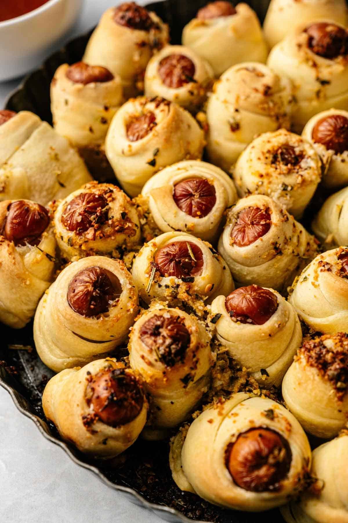 A tray of Pull-Apart Pigs in a Blanket, featuring mini sausages wrapped in golden-brown dough, sprinkled with herbs and grated cheese for a deliciously shareable treat.
