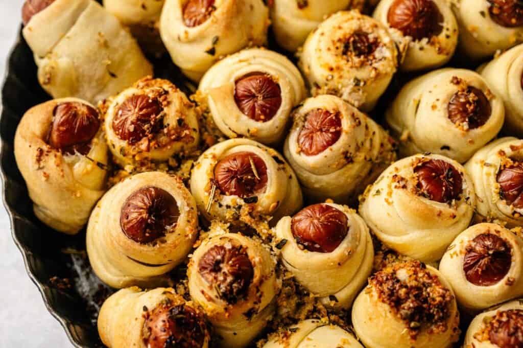 Pull-Apart Pigs in a Blanket, featuring mini sausages wrapped in golden pastry and sprinkled with herbs and seasoning, are arranged closely together on a tray for easy sharing.