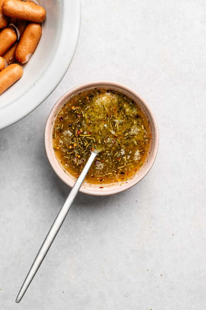 A bowl of seasoned dipping sauce with a spoon sits next to a plate of Pull-Apart Pigs in a Blanket mini sausages on a light surface.