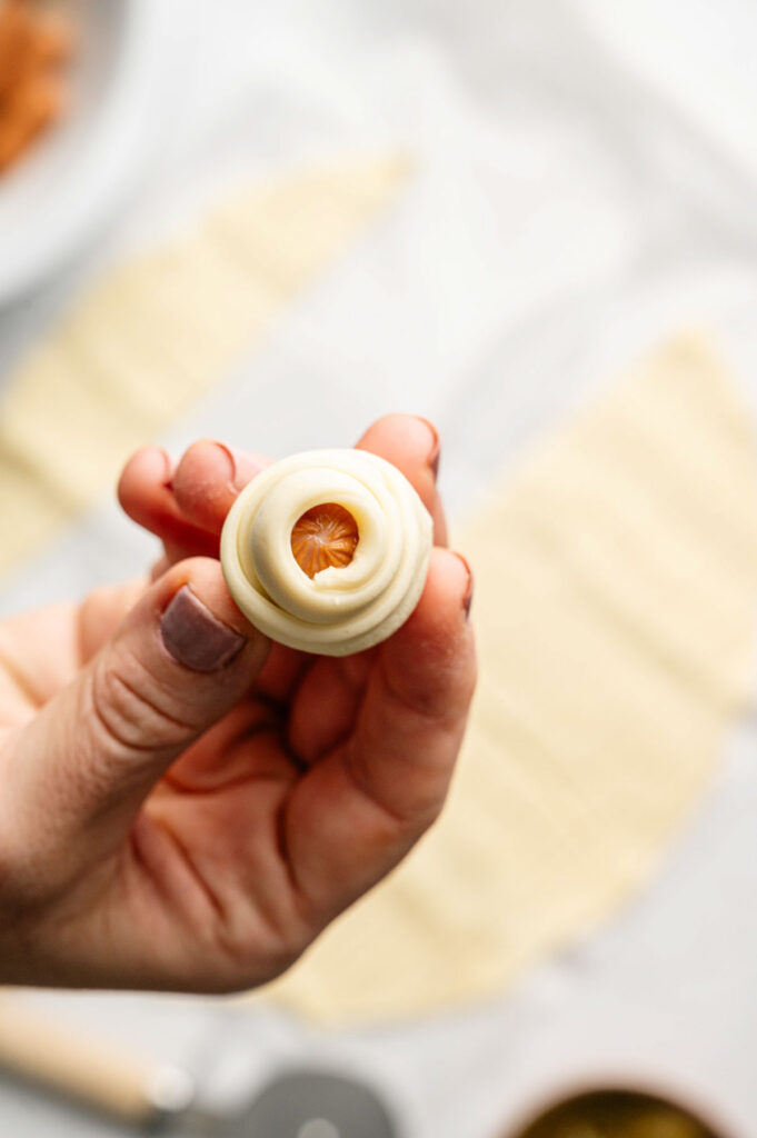 A hand holding a mini sausage wrapped and rolled in dough, showing the pinwheel technique before baking.