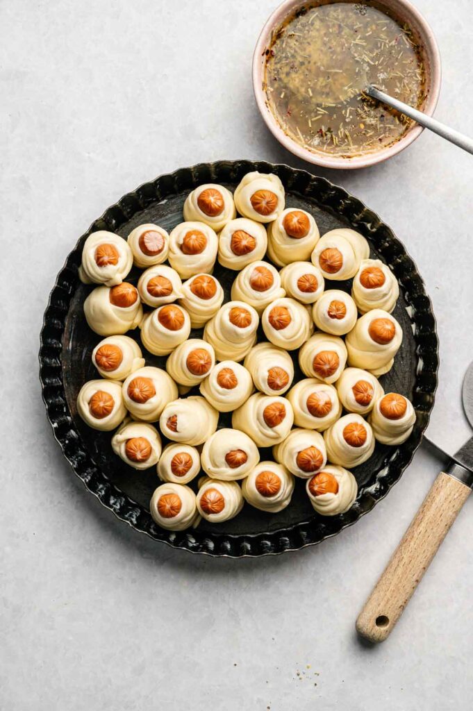 Unbaked Pull-Apart Pigs in a Blanket arranged in a circular pan, with a bowl of seasoning mixture and a pastry brush nearby on a light surface.
