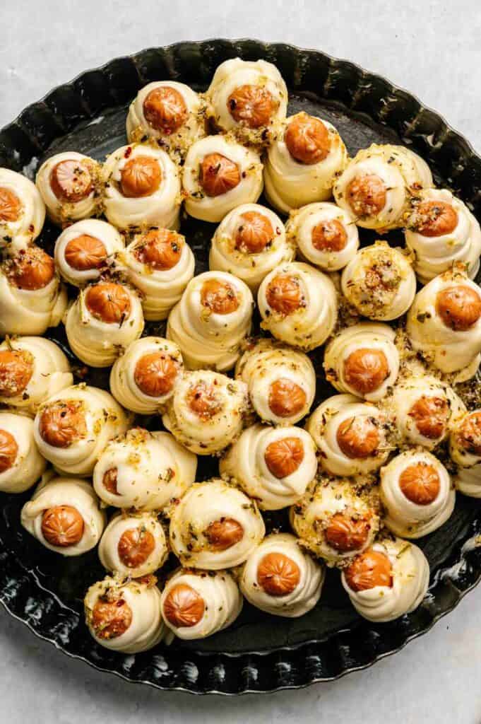 A round black tray filled with Pull-Apart Pigs in a Blanket&mdash;swirled pastry dough topped with small sausages and sprinkled with herbs and seasoning.