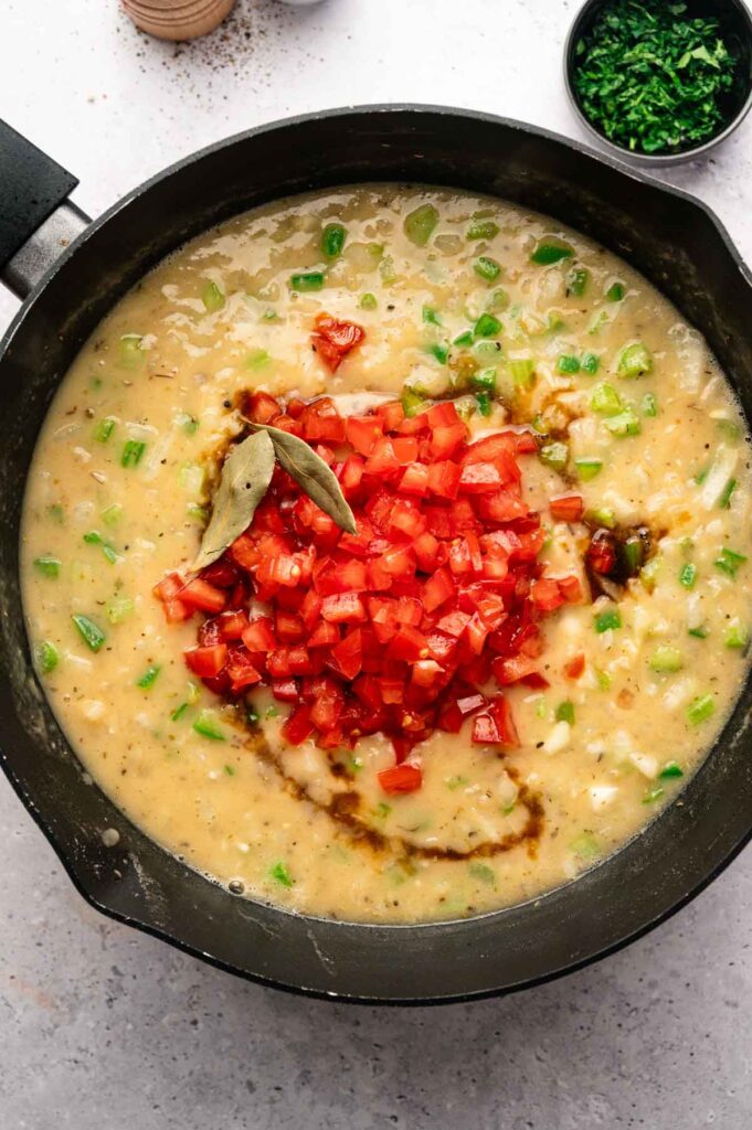 Diced tomatoes and bay leaves added to a simmering etouffee sauce base in a skillet.