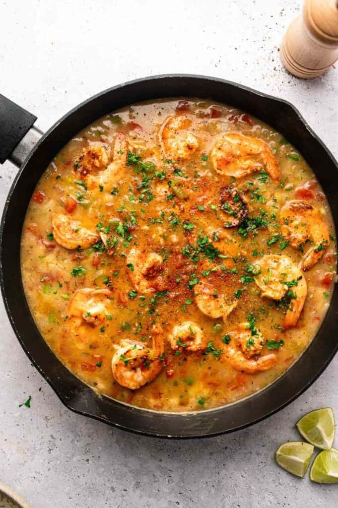 A skillet filled with creamy Shrimp Etouffee in a seasoned sauce, garnished with chopped herbs, with lime wedges and a pepper grinder nearby.