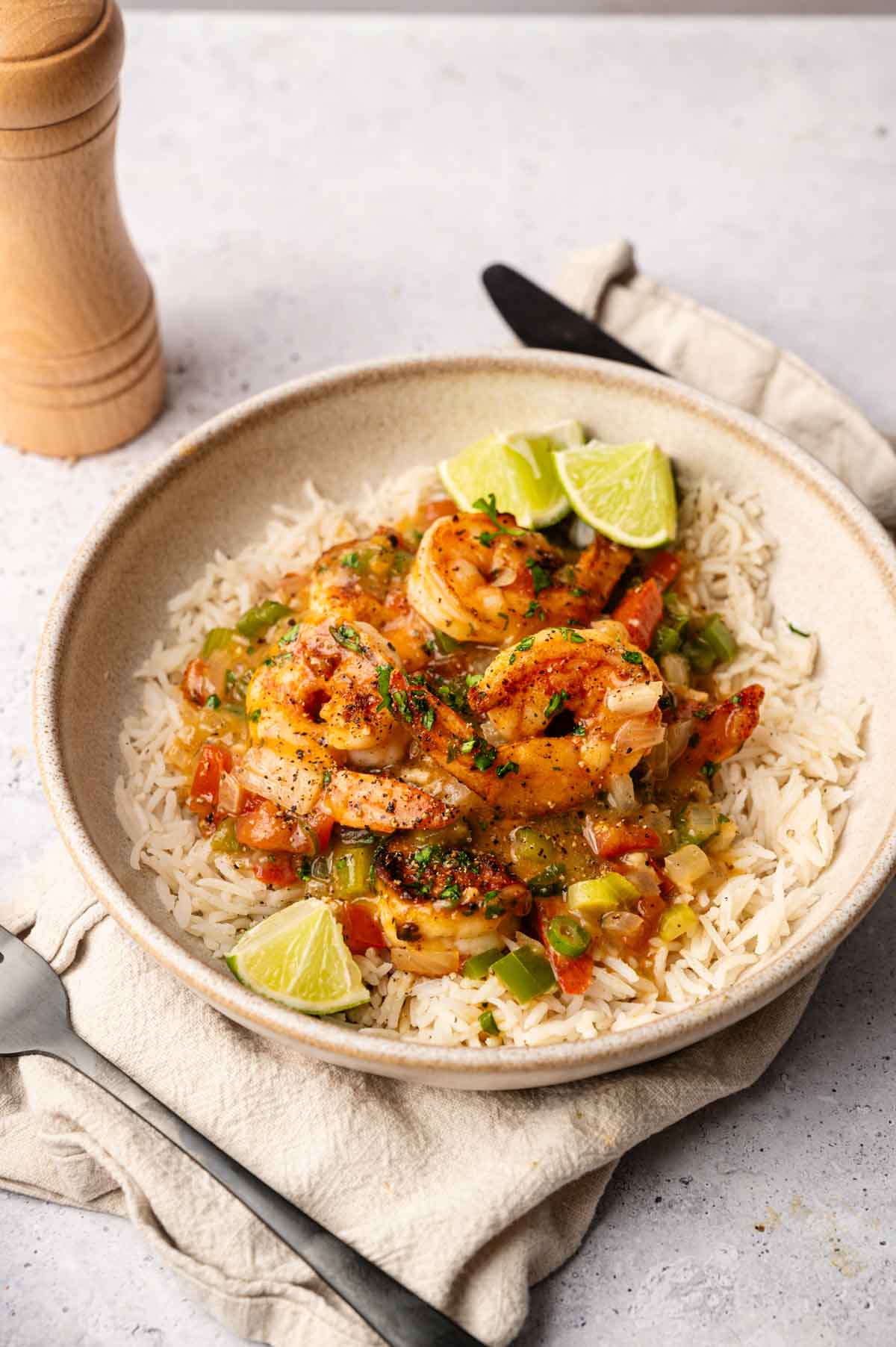 A bowl of white rice topped with cooked shrimp, diced vegetables, herbs, and lime wedges evokes the flavors of Shrimp Etouffee, set on a beige napkin with a fork and knife nearby.
