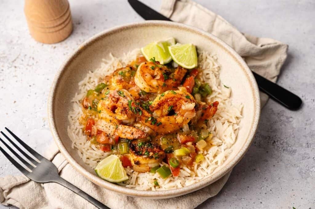 A bowl of white rice topped with Shrimp Etouffee, diced vegetables, and sauce, garnished with lime wedges, sits on a napkin next to a fork and knife.