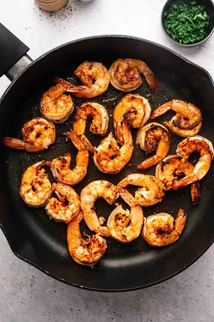Cooked, seasoned shrimp arranged in a single layer in a black skillet evoke the flavors of Shrimp Etouffee, with a bowl of chopped herbs visible in the background.