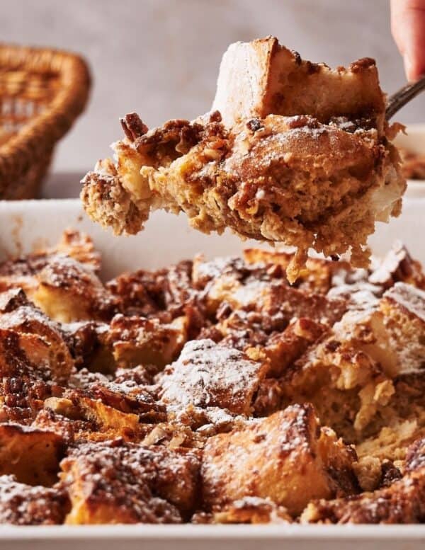 A hand serves a portion of bread pudding topped with powdered sugar and nuts from a baking dish.