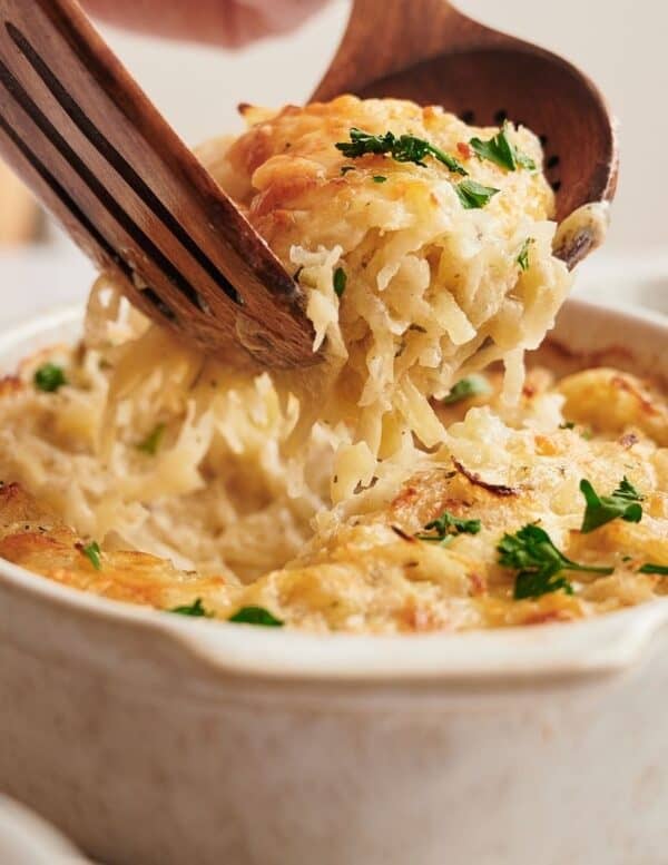 A wooden spoon serves a portion of cheesy baked casserole with shredded potatoes and parsley from a ceramic dish.