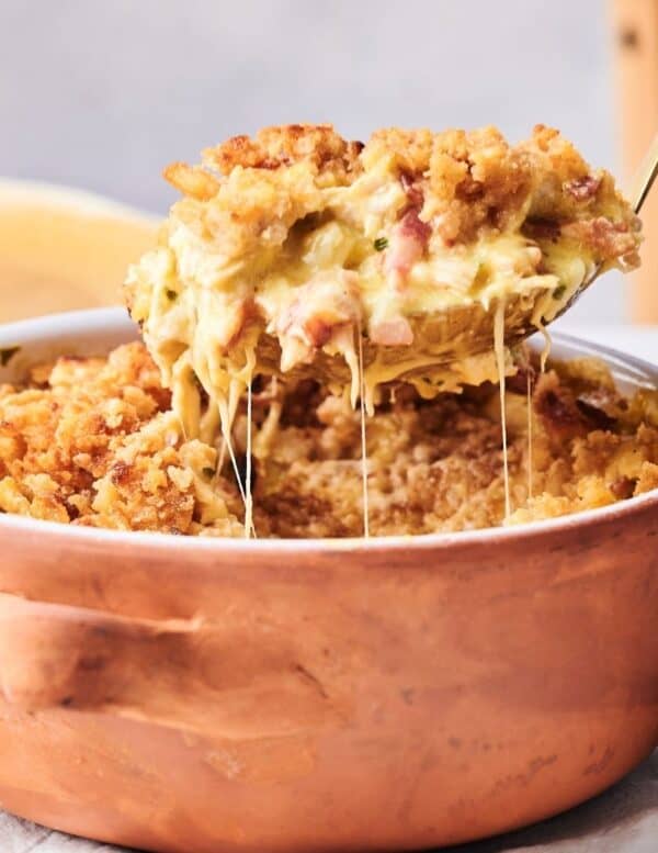 A spoon scoops out a portion of cheesy casserole with a breadcrumb topping from a copper-colored baking dish.