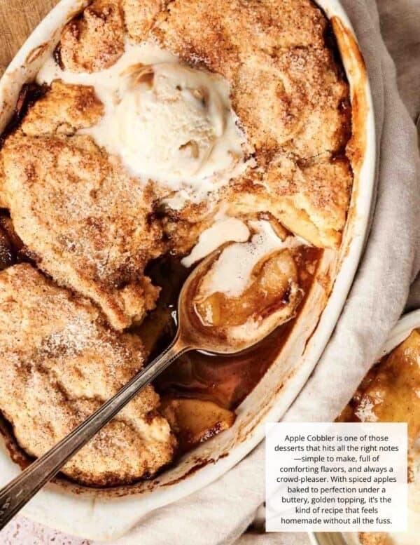 A baked apple cobbler in a white dish with a golden biscuit topping, a scoop of vanilla ice cream, and a serving spoon.