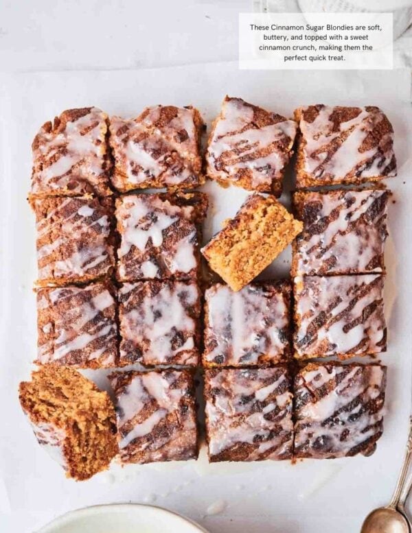 A tray of cinnamon sugar blondies cut into squares, topped with white icing drizzle, with one blondie turned to show the inside.