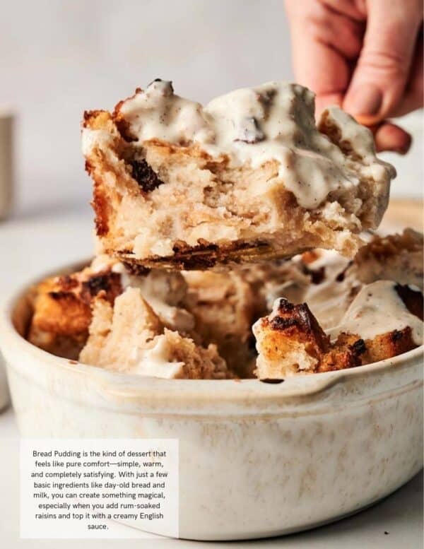 A serving spoon lifts a piece of bread pudding with raisins and creamy sauce from a white baking dish filled with more bread pudding.