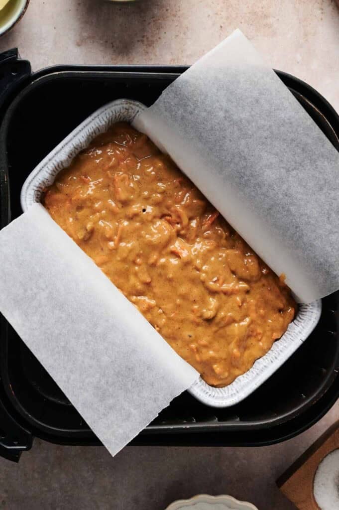 A loaf pan lined with parchment paper is filled with uncooked Air Fryer Carrot Cake batter and placed inside an air fryer.