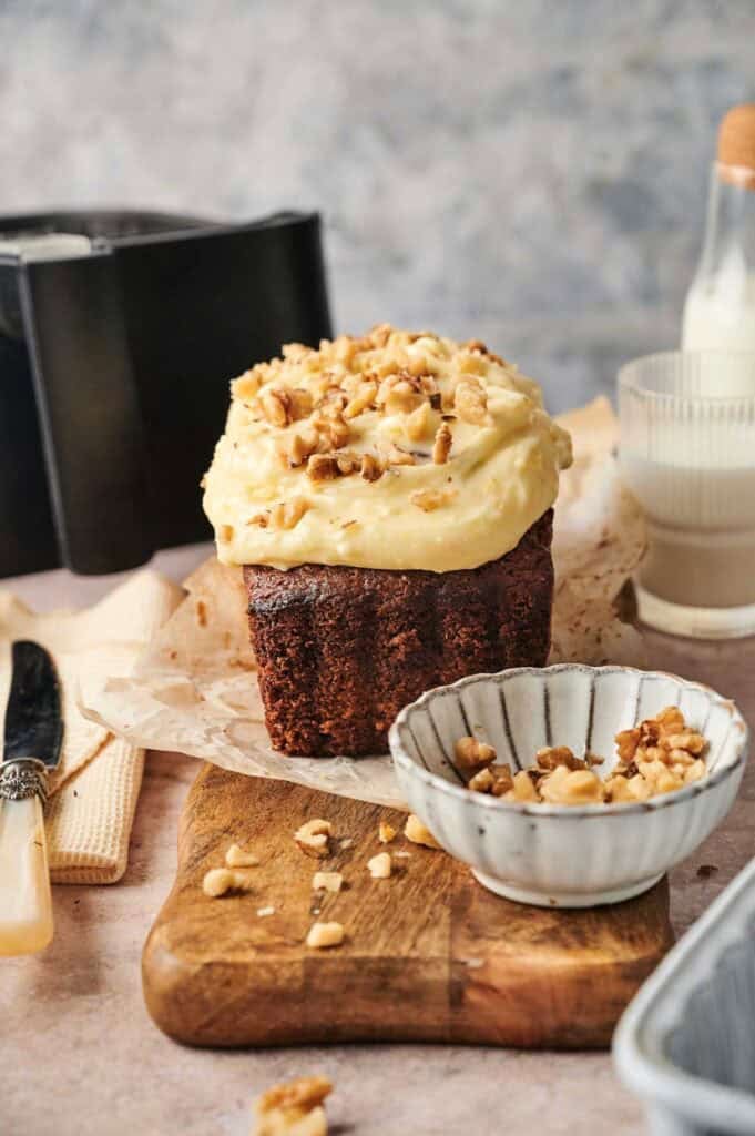 An Air Fryer Carrot Cake loaf with frosting and chopped walnuts on top sits on parchment paper on a wooden board, with a bowl of walnuts and a glass of milk nearby.
