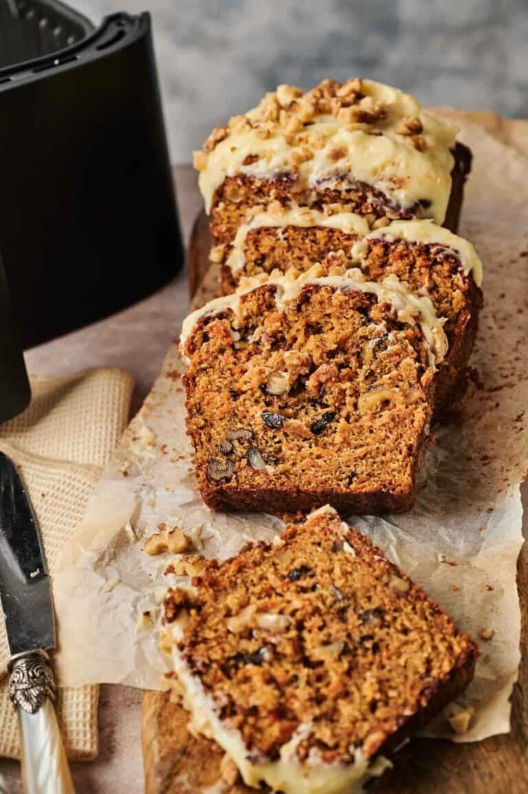 A sliced loaf of walnut and raisin bread with icing on top, displayed on parchment paper next to a knife and an Air Fryer Carrot Cake.
