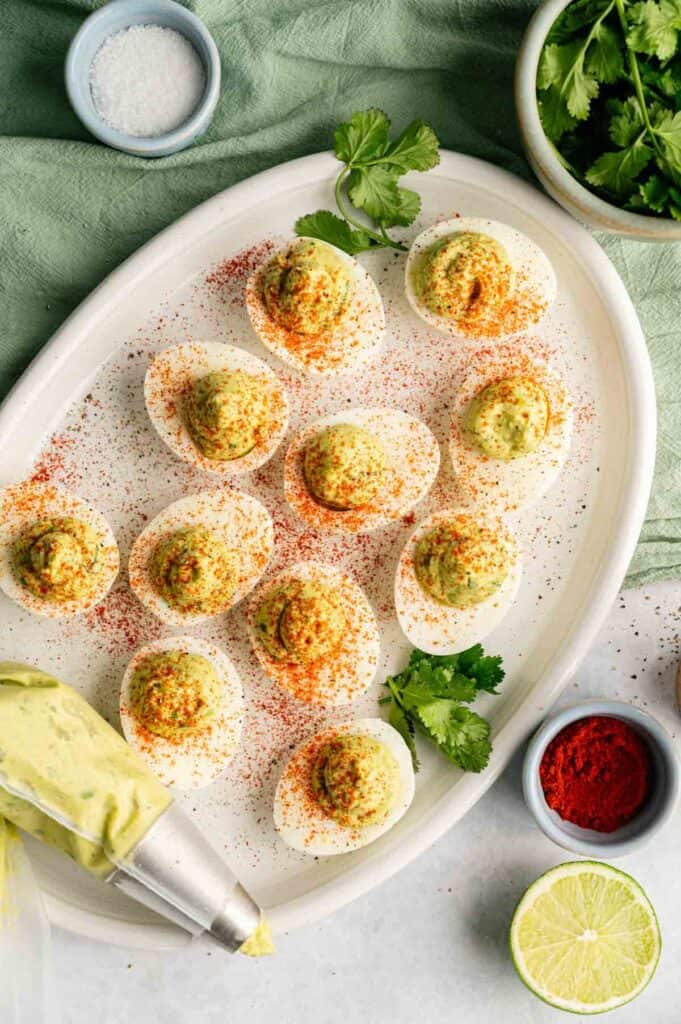 A plate of Avocado Deviled Eggs garnished with paprika and fresh herbs, surrounded by bowls of salt, paprika, cilantro, and a halved lime. A piping bag is visible in the corner.