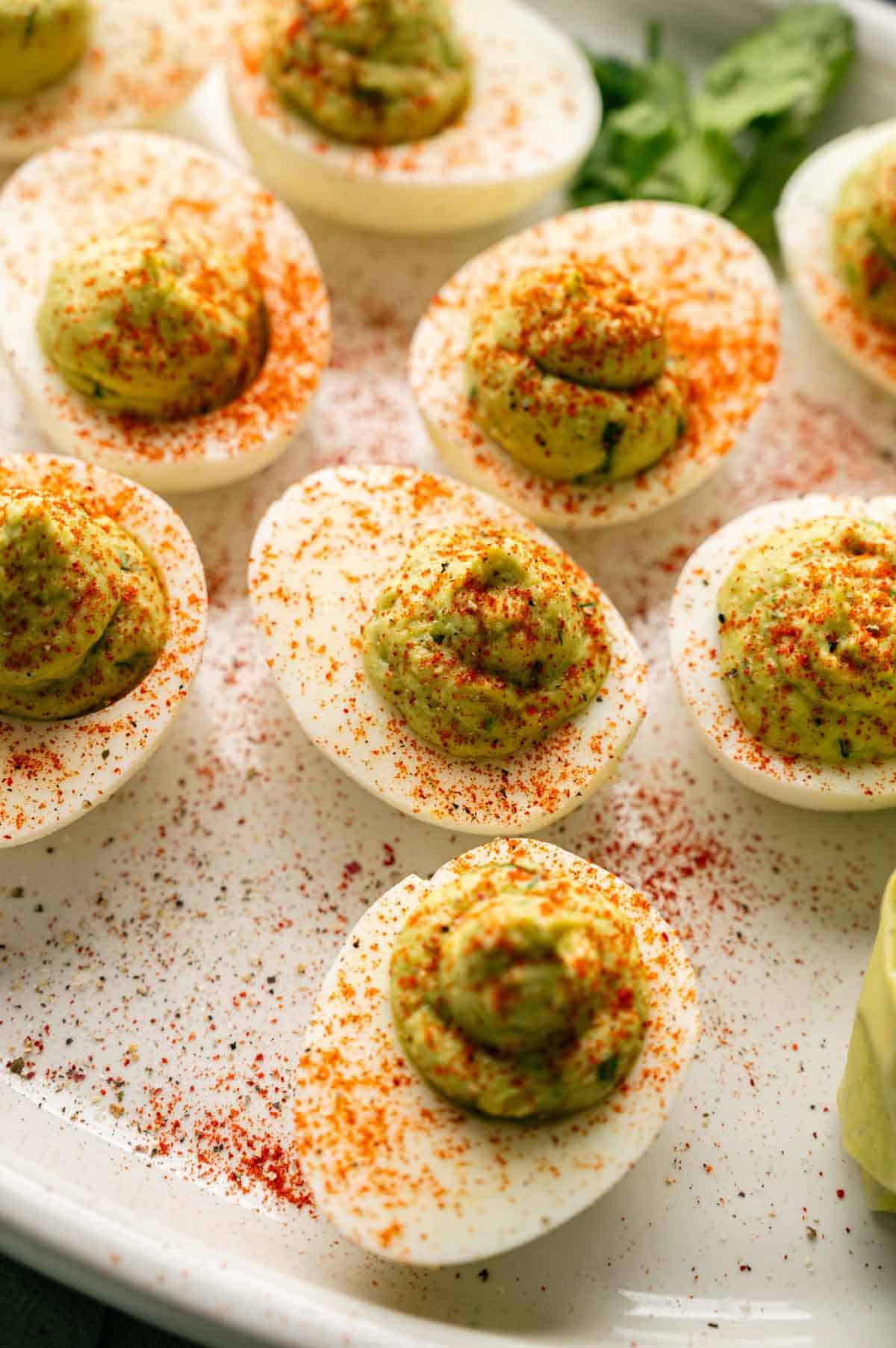 A plate of Avocado Deviled Eggs, topped with a creamy green filling and sprinkled with paprika, arranged artfully on a white dish.